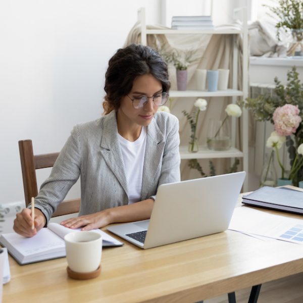 Planning work. Busy young female tailor flower arranger getting orders online in atelier domestic studio. Woman clothing designer sit by pc watch professional video course make notes in paper planner